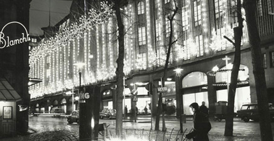 NK-huset mot Hamngatan i skymningsljus, gatubelysning och med juldekorationer. Cirka 1950–1970. NK:s historiska arkiv hos Centrum för Näringslivshistoria.